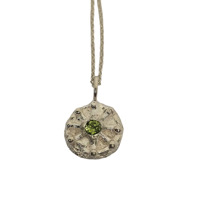 The Solar Plexus Pendant Peridot by Caroline Stokesberry-lee is shown on a chain, featuring a textured circular design with a green peridot gemstone at the center and small raised dots around the edge, displayed against a white background.