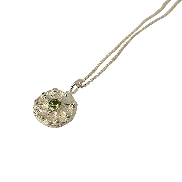 The Solar Plexus Pendant Peridot by Caroline Stokesberry-lee is a silver pendant necklace featuring a round, textured medallion set with a vibrant green peridot gemstone at its center, hand carved and displayed on a fine silver chain against a white background.