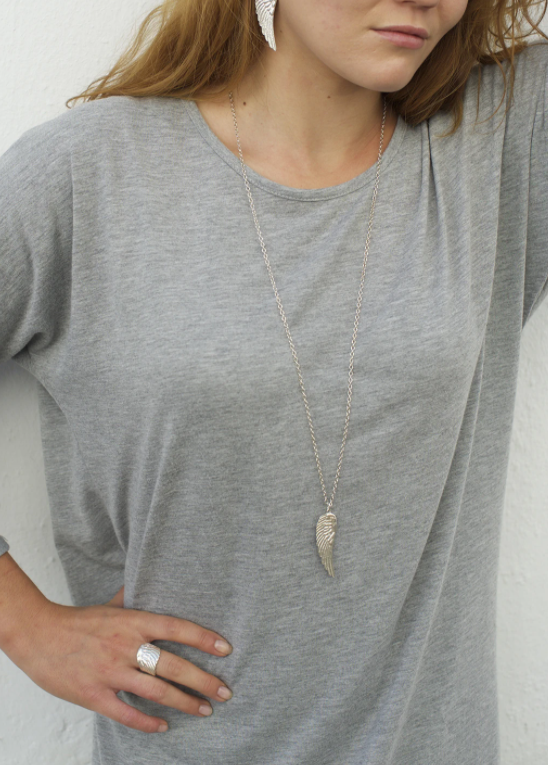 A person with long hair is wearing a light gray t-shirt paired with sterling silver jewelry, including Jennifer Kinnear's Angel Wing Necklace (silver & gold) and matching angel wing earrings, along with a large ring. The person stands with one hand resting on their hip.