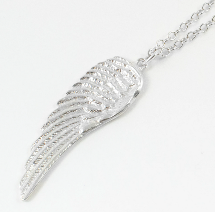 The Jennifer Kinnear Angel Wing Necklace features a delicate silver chain and a pendant shaped like an angel wing, intricately detailed with etched lines to resemble feathers, set against a simple white background.