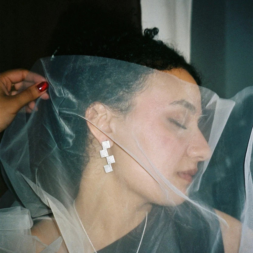 With eyes closed, a person wears Soufana Attia's Stairway to Heaven Earrings - Large and a sheer veil. A red-nailed hand gently lifts the veil, adding asymmetry and evoking a soft, dreamy mood.