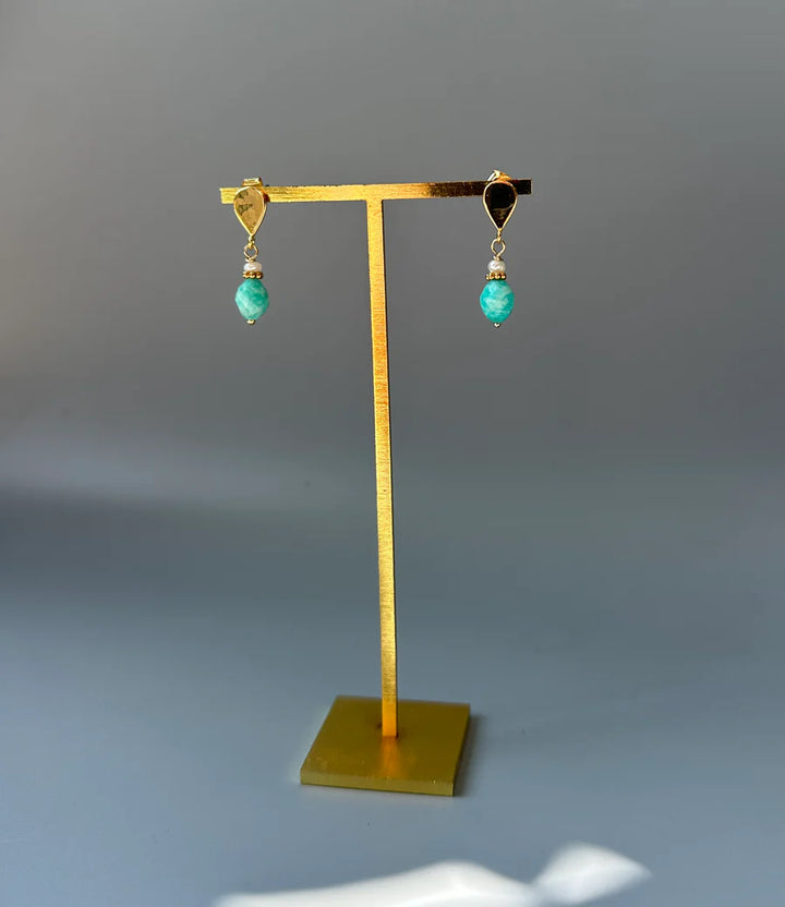 The Stanley Earrings by Aria V, featuring gold plating and teardrop turquoise stones, hang from a gold stand against a plain gray background.