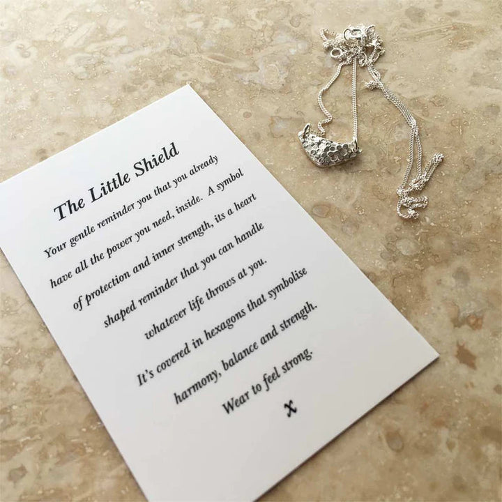 A Little Shield Necklace Silver by Andrea Mears Jewellery with a pendant in the shape of a small shield lies on a stone surface next to a card titled "The Little Shield." The card describes the pendant as a symbol of protection, inner strength, harmony, balance, and the power within.