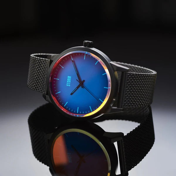 A Storm Styro Slate Blue wristwatch with a black mesh band is shown against a dark background. The watch face, featuring photochromic glass, has a blue gradient transitioning to orange and red at the edges, with slim, minimalist hour, minute, and second hands. The brand name "Storm Watches" is visible on the dial.