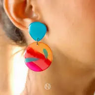 Close-up of a person wearing colorful, unique earrings. The top part is a blue teardrop shape, while the larger bottom part is a round disc with a vibrant abstract pattern in shades of orange, red, blue, and pink. These hand-painted Sunset Surf Statement Earrings by Naoi feature stainless steel earring posts for durability.