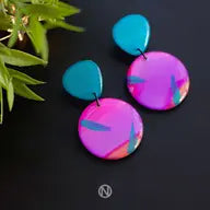 Unique, hand-painted Sunset Surf Statement Earrings by Naoi are displayed against a black background. The top part is a turquoise geometric shape, while the lower part is a larger circular piece in pink, purple, and orange with abstract designs. Green leaves are partially visible in the upper left corner.