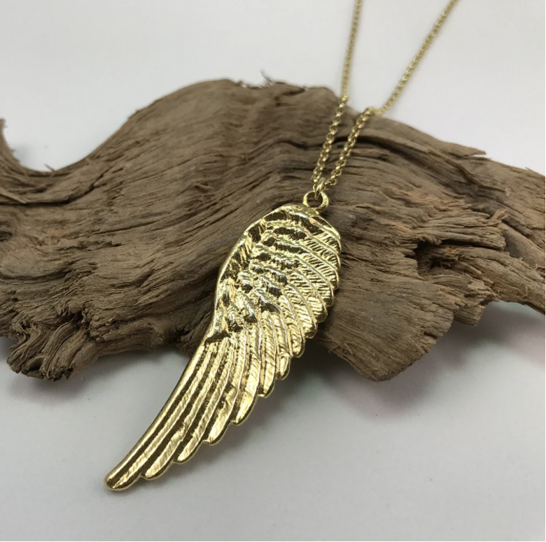 A Jennifer Kinnear Angel Wing Necklace (silver & gold) featuring a finely detailed feather design pendant on a delicate chain is laid on a piece of rough, textured driftwood. The exquisite craftsmanship of the pendant beautifully contrasts with the natural background.