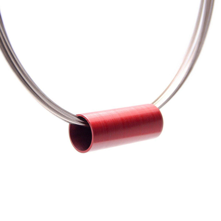 A modern statement necklace featuring a sleek, brushed red cylindrical pendant suspended on multiple thin, intertwined stainless steel wires. The design is minimalist and elegant, with the Tensor Tube Pendant by Filip Vanas's vibrant red color contrasting against the metallic sheen of the necklet.