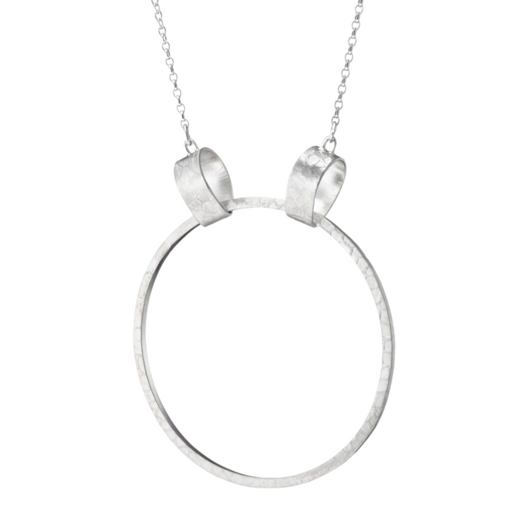 A minimalist sterling silver neckpiece, the Talún Ancaire Neckpiece Silver by Lynsey de Burca, features a thin, hammered metal hoop pendant suspended from a delicate chain. The chain loops through two small cylindrical connectors at the top of the hoop, adding a simple yet elegant touch to the design.