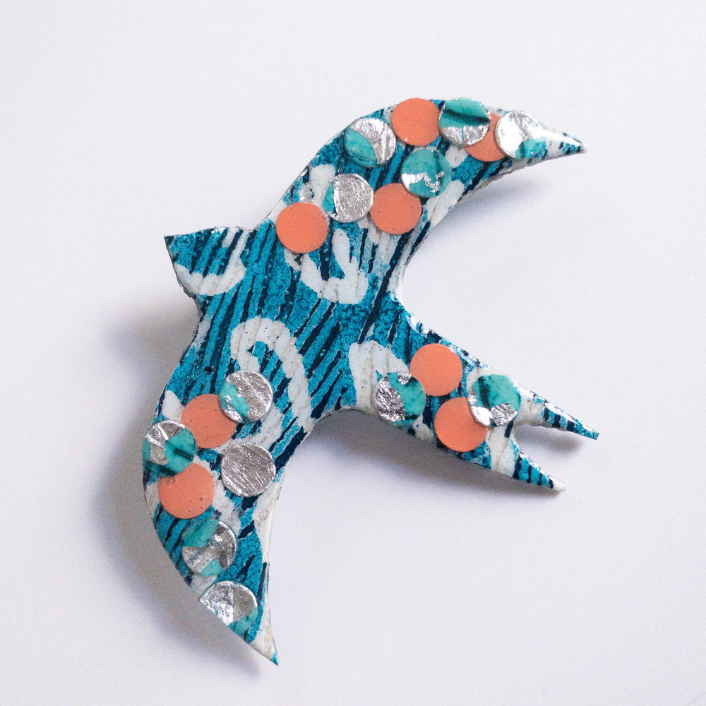 A handmade Éan Bird Batik Textile Brooch by Rothlú with a blue and white pattern, adorned with small orange and silver circular sequins. The bird's wings and tail are spread outward, creating a sense of motion. Its unique texture is set against a plain white background, highlighting the eco resin finish.