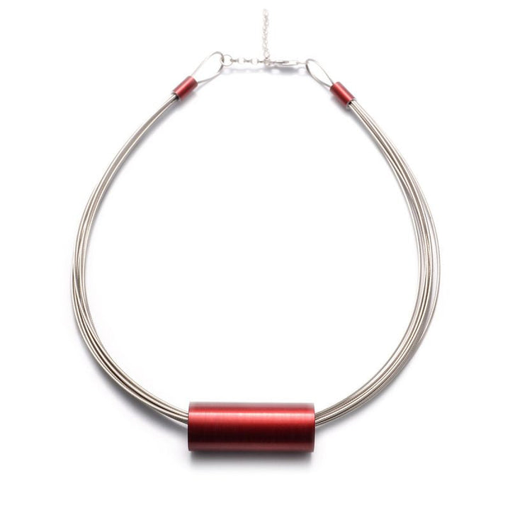 A sleek statement necklace featuring a thick, cylindrical red pendant threaded through multiple thin, metallic strands of stainless steel. The anodised aluminium clasp is visible at the top. The Tensor Tube Pendant by Filip Vanas has a design that is modern and minimalistic.