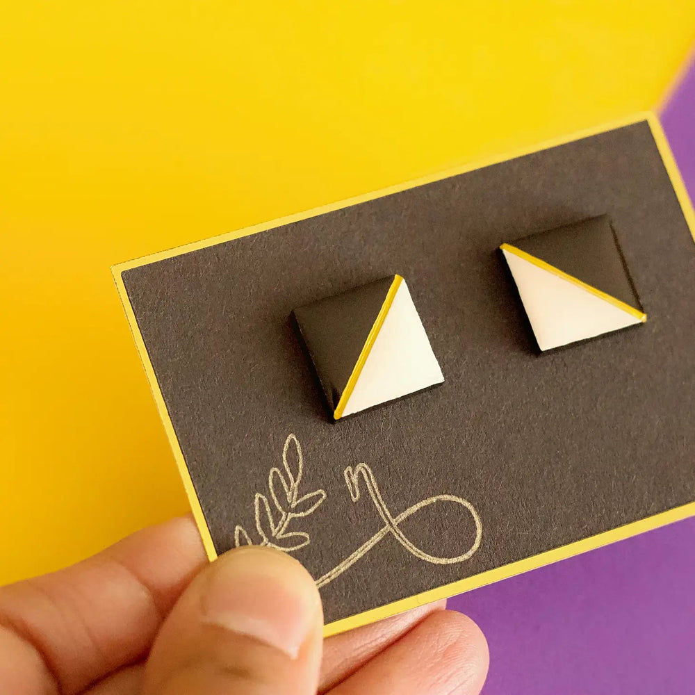 Close-up of a hand holding a card with Tetra Stud Earrings by Naoi. The card features a simple line drawing of a plant and is set against a vibrant yellow and purple background.