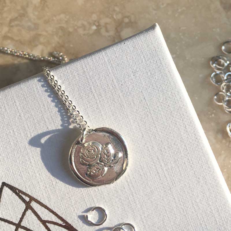 A sterling silver disc necklace with a round pendant featuring an embossed floral design, capturing the timeless symbol of love. The **Irish Rose Necklace Silver by Andrea Mears Jewellery**, highlighting an Irish Rose motif, is displayed on a textured white surface, catching light and casting a shadow.