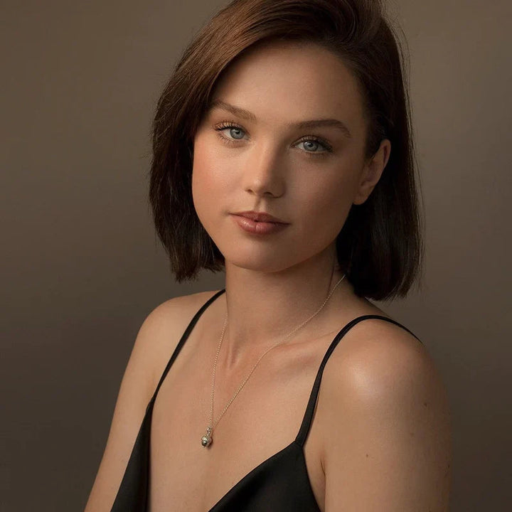 A woman with straight, shoulder-length dark brown hair is wearing a simple black spaghetti strap top and a sterling silver necklace with a small acorn pendant. She is looking directly at the camera with a neutral expression against a plain brown background. The Little Acorn Necklace Gold by Andrea Mears Jewellery adds an elegant touch to her outfit.