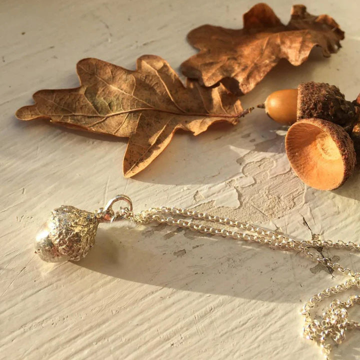 A The Little Acorn Necklace Gold by Andrea Mears Jewellery is laid on a white surface, with dried autumn leaves and an acorn cap as decoration. Sunlight casts soft shadows, adding warmth to the scene.