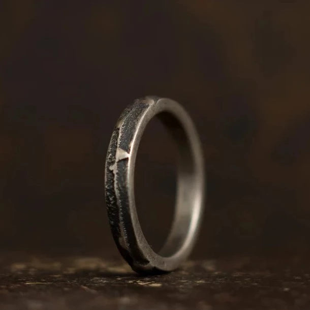 A close-up of a rustic, textured metal ring standing upright against a dark, blurred background. The Thin Ribbon Ring by Septentrion, made from patinated 925 silver, has a rough, distressed surface with visible imperfections, giving it a rugged and handmade jewellery appearance. The lighting casts subtle shadows on the unique variations of the ring.