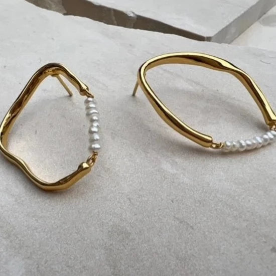 A pair of Thiva Earrings by Shyla, uniquely shaped in 22-carat gold, rests on a stone surface. Each features a wavy, irregular gold loop adorned with small white pearls. The design gives a modern, artistic touch to traditional jewelry elements.