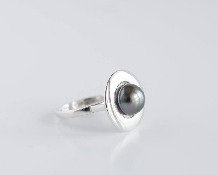 A sterling silver ring featuring a large, round, Tahitian Pearl set in a wide, circular bezel on a plain, sleek band. The design is minimalistic and elegant, contrasting the dark pearl with the shiny silver setting, presented against a plain white background. This exquisite piece is known as the Home Planet Silver Tahitian Pearl by Cosmic Boulevard.