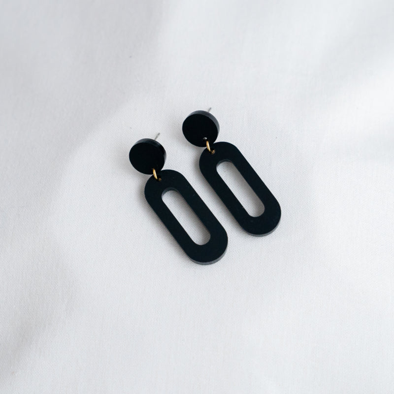 A pair of Timeless 04 Oval Acrylic Earrings by Studio Nok Nok is displayed on a white fabric background. Each earring features a small circular stud connected to a larger elongated oval-shaped hoop, creating a minimalist and modern design—perfect earrings that embody the essence of handmade jewelry.