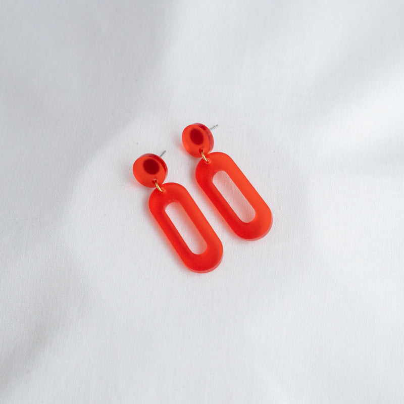 A pair of vibrant red Timeless 04 Oval Acrylic Earrings by Studio Nok Nok lies on a white surface. These handmade jewelry pieces feature a simple geometric design with a small circular stud connected to a larger, elongated oval-shaped drop. The minimalist design is both modern and bold, making them the perfect earrings for any occasion.