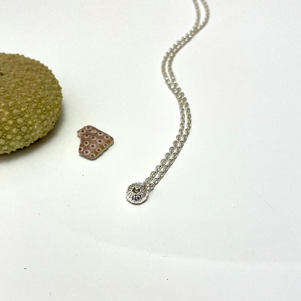 The Sea Urchin Necklace Tiny by Jennifer Kinnear, in gold plated silver, is shown on a white surface beside a green sea urchin shell and a small shard of pink patterned pottery.
