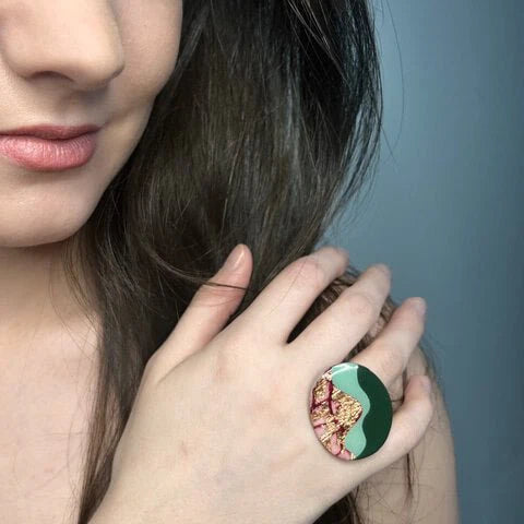 A woman with long brown hair wears the Rothlú Tonn Ring in Red, Rose-Gold, Mint, and Olive on her index finger as she touches her face and smiles slightly.