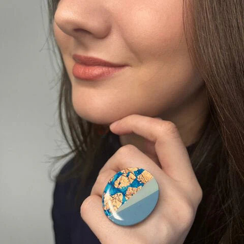 A woman with light skin and brown hair smiles gently, her chin resting on her hand, wearing the Rothlú Tonn Ring in Turquoise/Blue/Rose Gold—an eye-catching piece of handcrafted textile jewelry—on her finger.