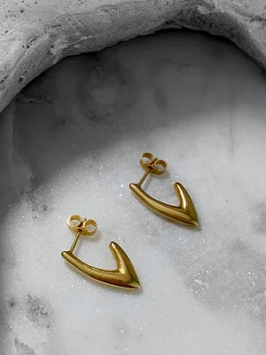 The Treo Mini Earrings by Inner Island, featuring a gold arrow-shaped minimalist design, are displayed on a smooth light gray stone with a textured edge—ideal for everyday wear or layering in your ear stack.