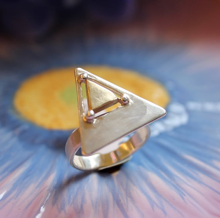 A close-up of the Triune Ring by Gallardo & Blaine Designs, crafted in sterling silver with a flat triangular shape and geometric details, set against a background of blue and yellow floral patterns.