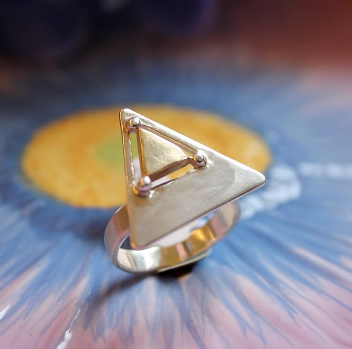 A close-up of the Triune Ring by Gallardo & Blaine Designs, crafted in sterling silver with a flat triangular shape and geometric details, set against a background of blue and yellow floral patterns.