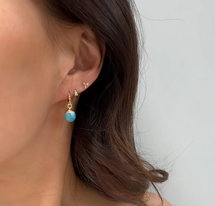 A close-up shows a woman's ear wearing Aria V’s handmade Turquoise Drop Earrings, a gold stud, and a large hoop with a faceted turquoise bead. Her brown hair is partially visible against a light background.