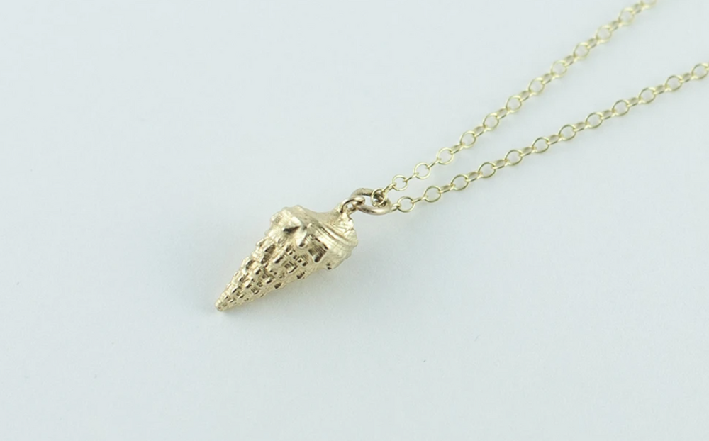 A delicate necklace with a gold-toned chain and a small pendant shaped like an ice cream cone. The pendant features intricate detailing, including the cone texture and a swirled ice cream scoop on top, reminiscent of lazy afternoons in Menorca. The background is a plain light grey color. Introducing the Whelk Shell Necklace by Jennifer Kinnear.