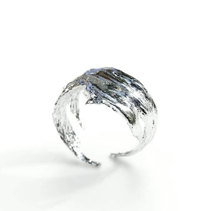 A wide, textured silver ring with an open end, appearing rugged and organic in design, much like a natural piece of bark. The Caroline Stokesberry-lee Woodland Bark Ring Silver has a polished, reflective surface with a unique, freeform shape and is positioned upright against a plain white background.