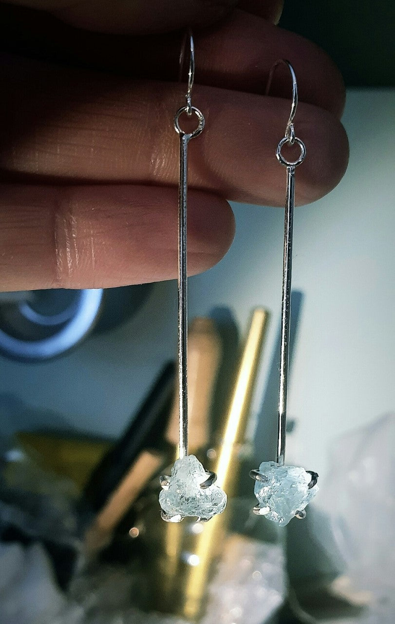 A hand holding a pair of Raw Aquamarine Drop Earrings by Caroline Stokesberry-lee with raw, light blue aquamarine gemstones at the bottom. The background is blurry, showcasing indistinct objects and tools. These earrings are delicate and minimalist in design, featuring elegant ear wires.