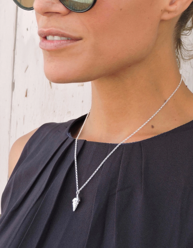 A close-up of a person wearing round sunglasses and a black sleeveless top, showcasing a delicate silver necklace with a small, pointed pendant. The background features light-colored walls reminiscent of the sunny ambiance of the Balearic Islands. The necklace is the Whelk Shell Necklace by Jennifer Kinnear.