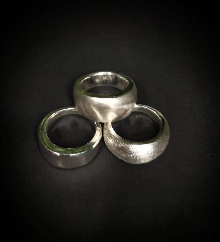 Three silver rings are arranged in a triangle formation on a black background. The rings have a polished, slightly reflective surface, and each has a unique texture: one is smooth, one is brushed, and one has a hammered finish. This composition highlights the expertise of jewellery artisans with the Silver Works - Ring Carving Kit by Silver Works.