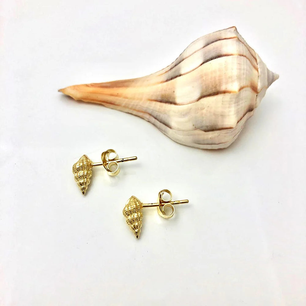 The Jennifer Kinnear Whelk Shell Stud Earrings in gold, inspired by Menorca shell jewelry, are shown on a white background beside a large natural seashell in pale brown and cream tones.