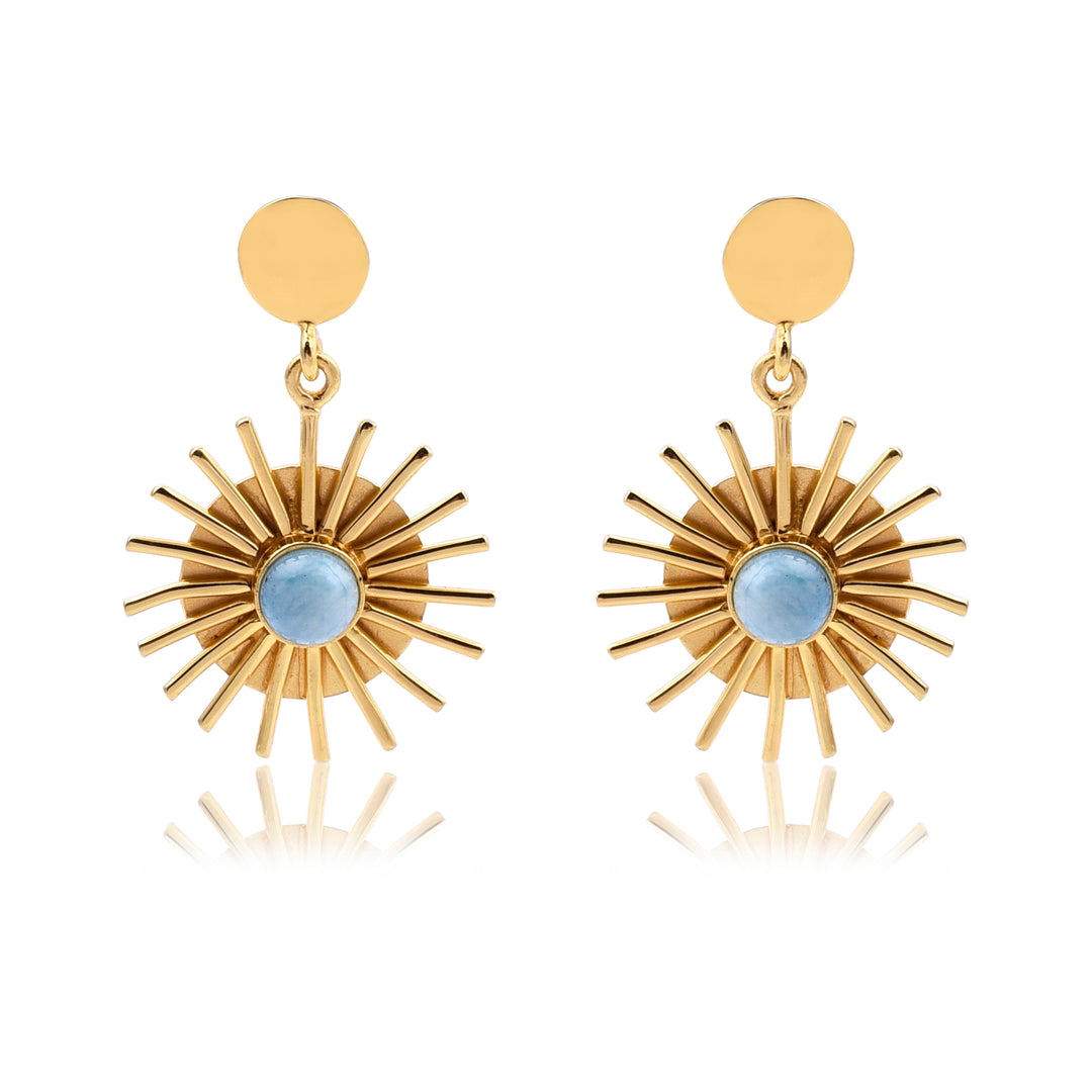 These earrings have a striking, radiant design that evokes the image of a sunburst. There's a small golden disc that sits in the earlobe—simple and smooth. From this disc hangs a larger, eye-catching design that looks like a miniature sun: a circular frame with golden rays extending outward in all directions. At the center of this sunburst is a round, sky-blue stone.