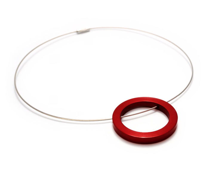 Introducing the Filip Vanas Circle Pendant in silver, a minimalist masterpiece. This necklace features a thin wire and showcases a striking red anodized aluminum pendant with an open center, giving it a distinctive doughnut shape. Its simple and modern design is sure to make a bold statement.