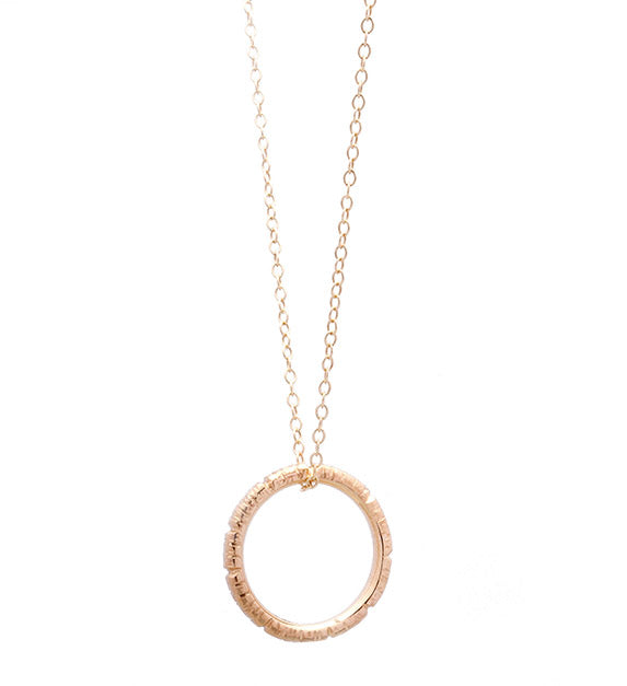 A delicate gold necklace features a thin chain with a circular pendant. The textured, uneven surface of the gold pendant lends it a rustic yet elegant appearance. This unique necklace offers minimalist and timeless charm, making it an exquisite piece of handmade jewelry. The Into The Light Circle Pendant 9ct Yellow Gold by Aud epitomizes these qualities beautifully.