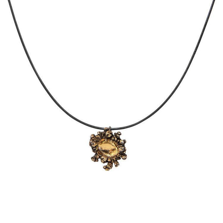 A black necklace features a statement pendant with 24K gold plating and an intricate design. The Spore Small Pendant Black & Gold by Abbie Dixon consists of a central, faceted amber-colored gemstone surrounded by multiple small beads and details forming a floral or sunburst pattern. The background is plain white.