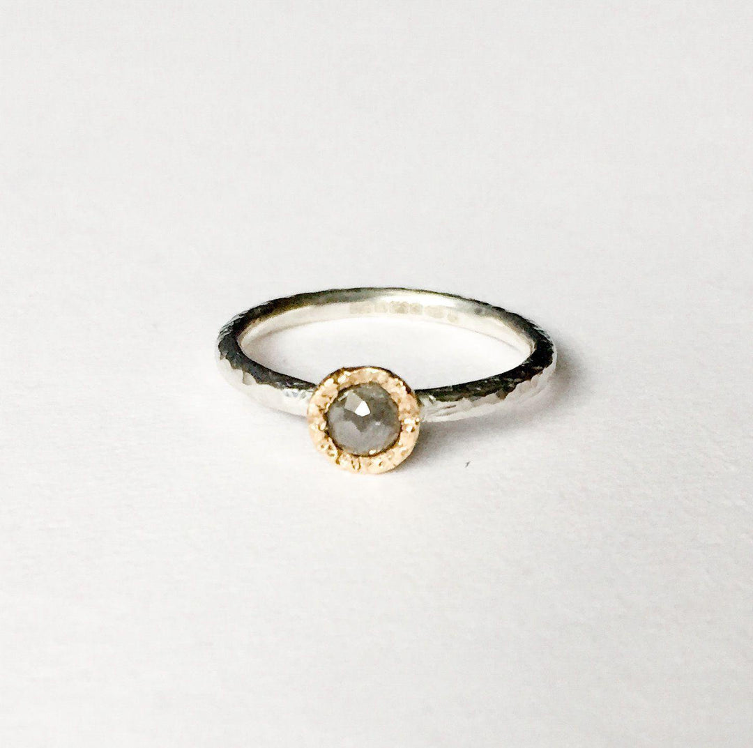 A minimalist ring featuring a single, round grey diamond set in a textured gold bezel on a slim, hammered finish sterling silver stacking ring. The Aurous Ring Silver Grey Diamond by Abbie Dixon is placed against a plain white background.