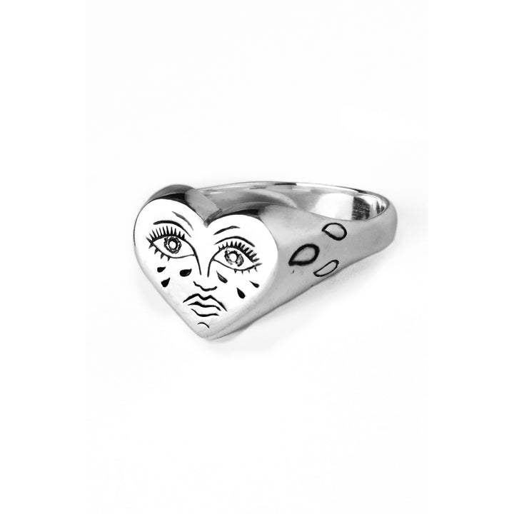 The Sad Lil Grimm Signet by Cosmic Boulevard features a silver ring with a heart-shaped face on the top, resembling a crying heart tattoo. The face is hand engraved with eyes, lashes, and tear-like markings around it, giving it a distinctive, artistic look. Handmade in Dublin, the band also features three small engraved shapes on the side against a white background.