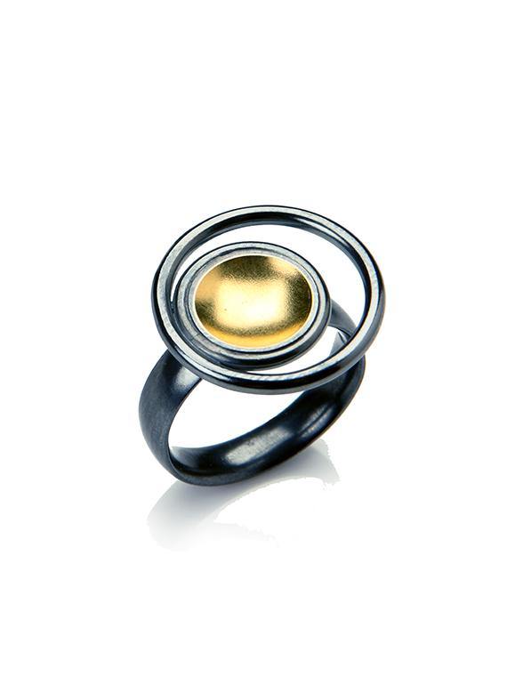 A modern, artistic ring features a brushed black band with a large, circular design at its top. The central disk is 22ct yellow gold and surrounded by an intricate pattern of oxidised silver and black concentric loops, creating a striking, contemporary aesthetic. The **Black & Gold Ring** by **Fiona Kerr** embodies this unique design.