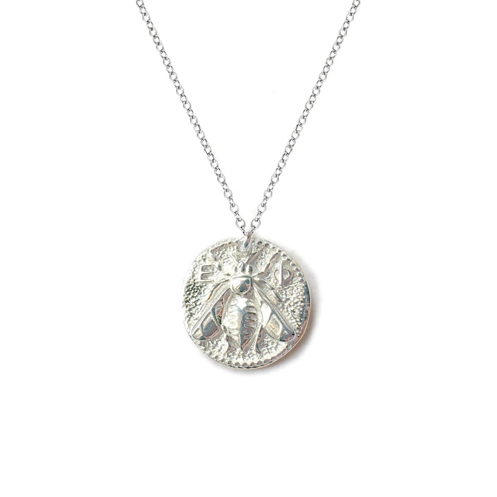 The Queen Bee Necklace Silver by Andrea Mears Jewellery is a sterling silver necklace featuring a round pendant with an intricately detailed bee design reminiscent of an ancient Greek coin. The queen bee appears in the center, surrounded by subtle texture accents on the pendant's surface. The piece is suspended from a delicate silver chain.