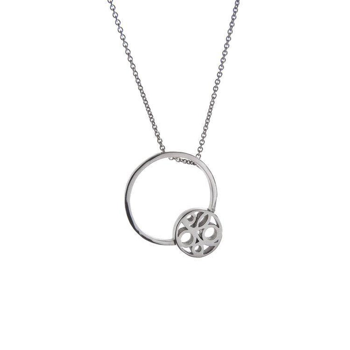 A Miriam Wade Flow Pendant Silver featuring two interconnected circular elements. One circle is plain, while the other showcases a decorative cutout pattern. The delicate and minimalistic necklace chain complements the intricate design. The background is plain white, enhancing its elegance.