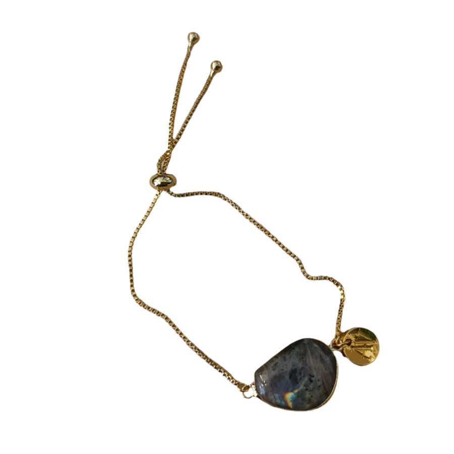 A delicate gold chain Ria Bracelet with an adjustable closure featuring a teardrop-shaped stone charm and a small round gold charm by Aria V. The teardrop-shaped stone has an iridescent quality, reflecting hues of green and blue. The bracelet is laid on a wooden surface.