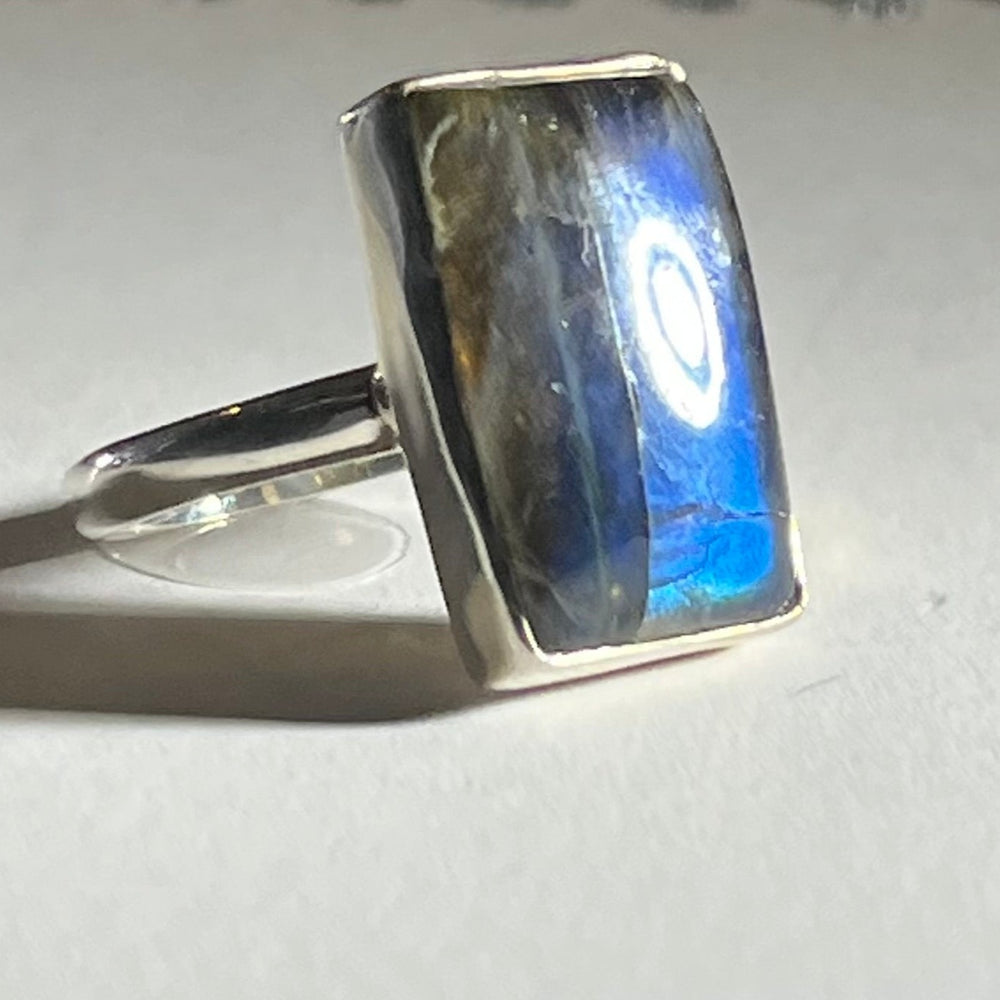 A **Rectangular Labradorite Ring** by **Watermelon Tropical** featuring a rectangular labradorite gemstone with vibrant blue and green iridescent hues. The stone's polished surface reflects light, creating a radiant shine. The ring is placed on a light-colored background.