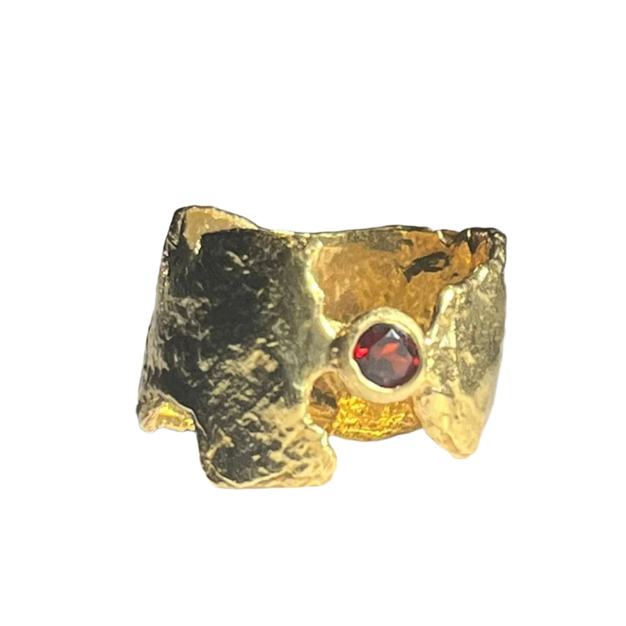 A rough-textured gold-plated ring with an uneven, organic shape is pictured on a white surface. The Faerie Tale Garnet Ring Gold Plate by Caroline Stokesberry-lee features a garnet set into one side, creating a striking contrast with the gold. Shadows are cast on the surface, highlighting the ring's rugged design.