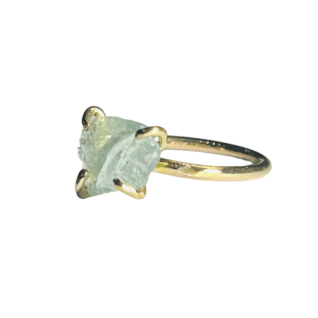 A Raw Aquamarine 14ct Gold Filled Ring by Caroline Stokesberry-lee with a rough, unpolished light green stone set in a minimalist, four-prong setting. The ring is placed on a smooth, light-grey surface, and dramatic lighting creates soft shadows, highlighting the texture and color of the stone.